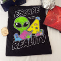 A black graphic t-shirt laid flat on a white fabric background, featuring a colorful design of a green alien reading a pink book with the words 'ESCAPE REALITY' in bold white letters. The shirt is styled with a small wooden sci-fi badge, a dark blue box, tin foil hats, and a book open to a solar system map.