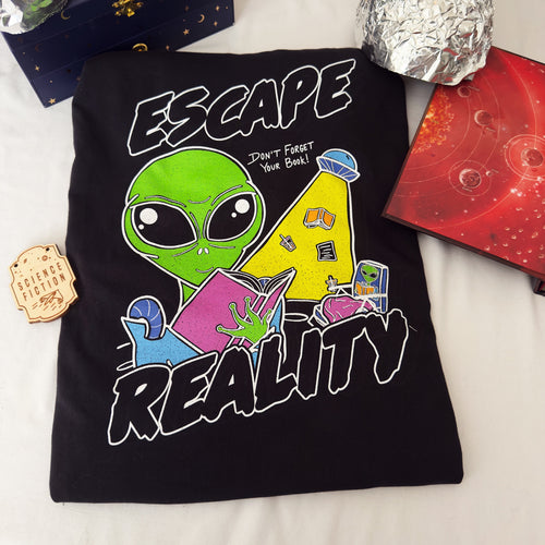 A black graphic t-shirt laid flat on a white fabric background, featuring a colorful design of a green alien reading a pink book with the words 'ESCAPE REALITY' in bold white letters. The shirt is styled with a small wooden sci-fi badge, a dark blue box, tin foil hats, and a book open to a solar system map.