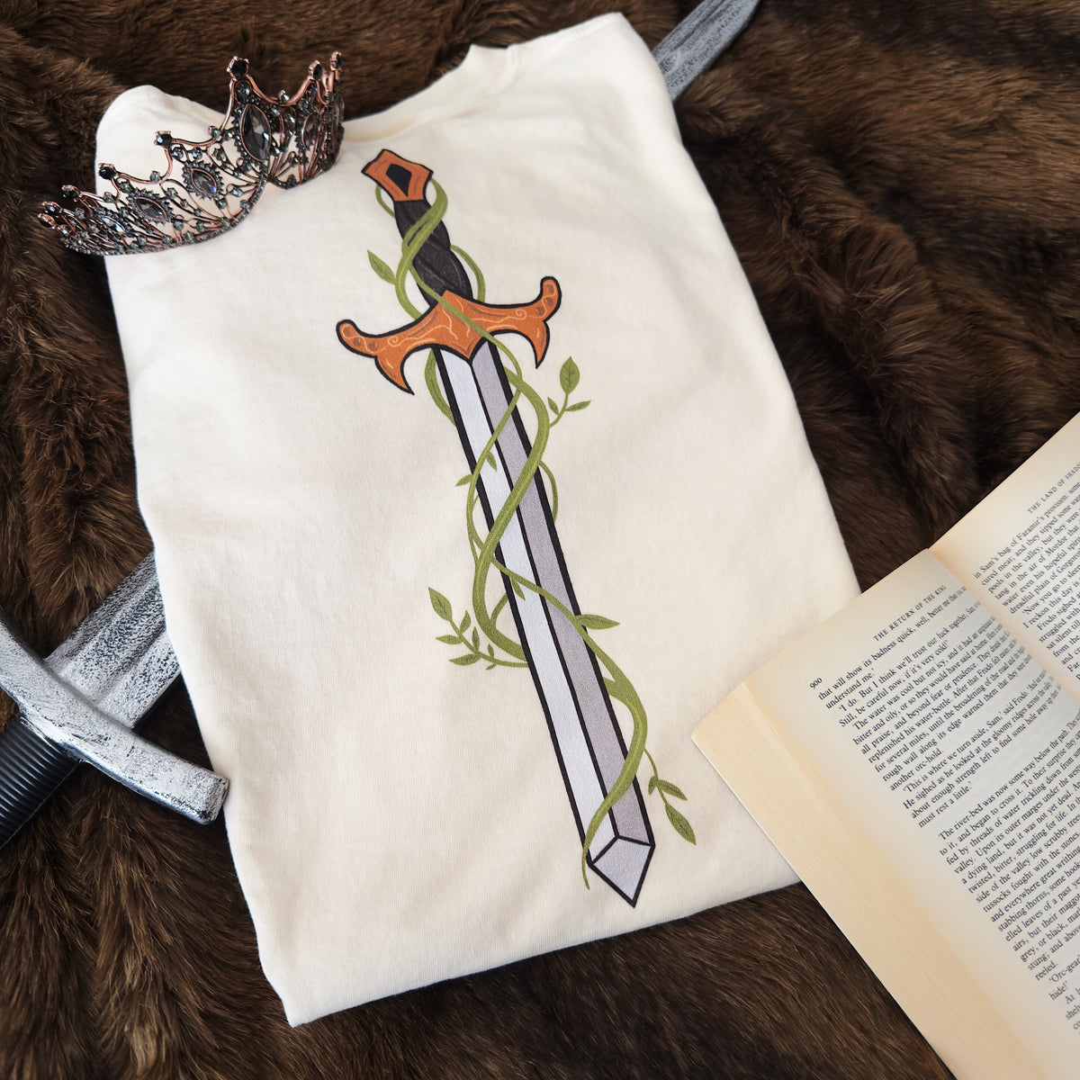 A cream t-shirt featuring a graphic of a sword wrapped in green vines, styled in a flat-lay with a crown, an open book, and a silver sword on a brown faux fur background.