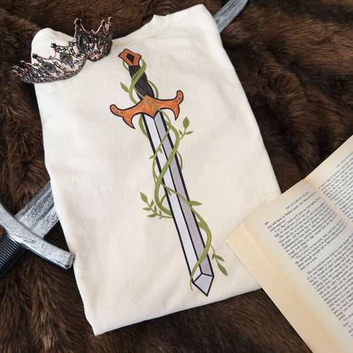 A cream t-shirt featuring a graphic of a sword wrapped in green vines, styled in a flat-lay with a crown, an open book, and a silver sword on a brown faux fur background.