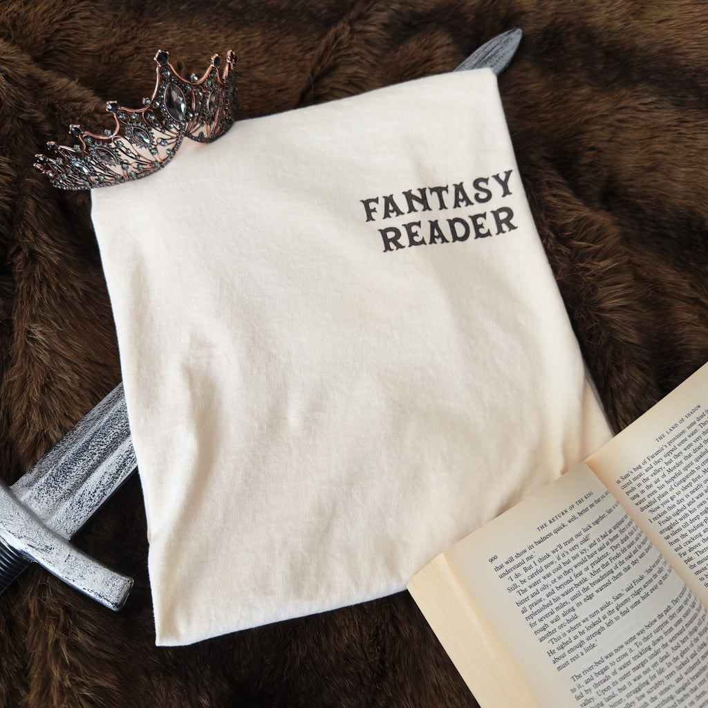 A cream-colored t-shirt printed with the words 'FANTASY READER' on the left chest in a dark brown serif font, styled on a brown faux fur background with a silver sword, a bronze crown, and an open book.
