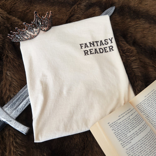 A cream-colored t-shirt printed with the words 'FANTASY READER' on the left chest in a dark brown serif font, styled on a brown faux fur background with a silver sword, a bronze crown, and an open book.