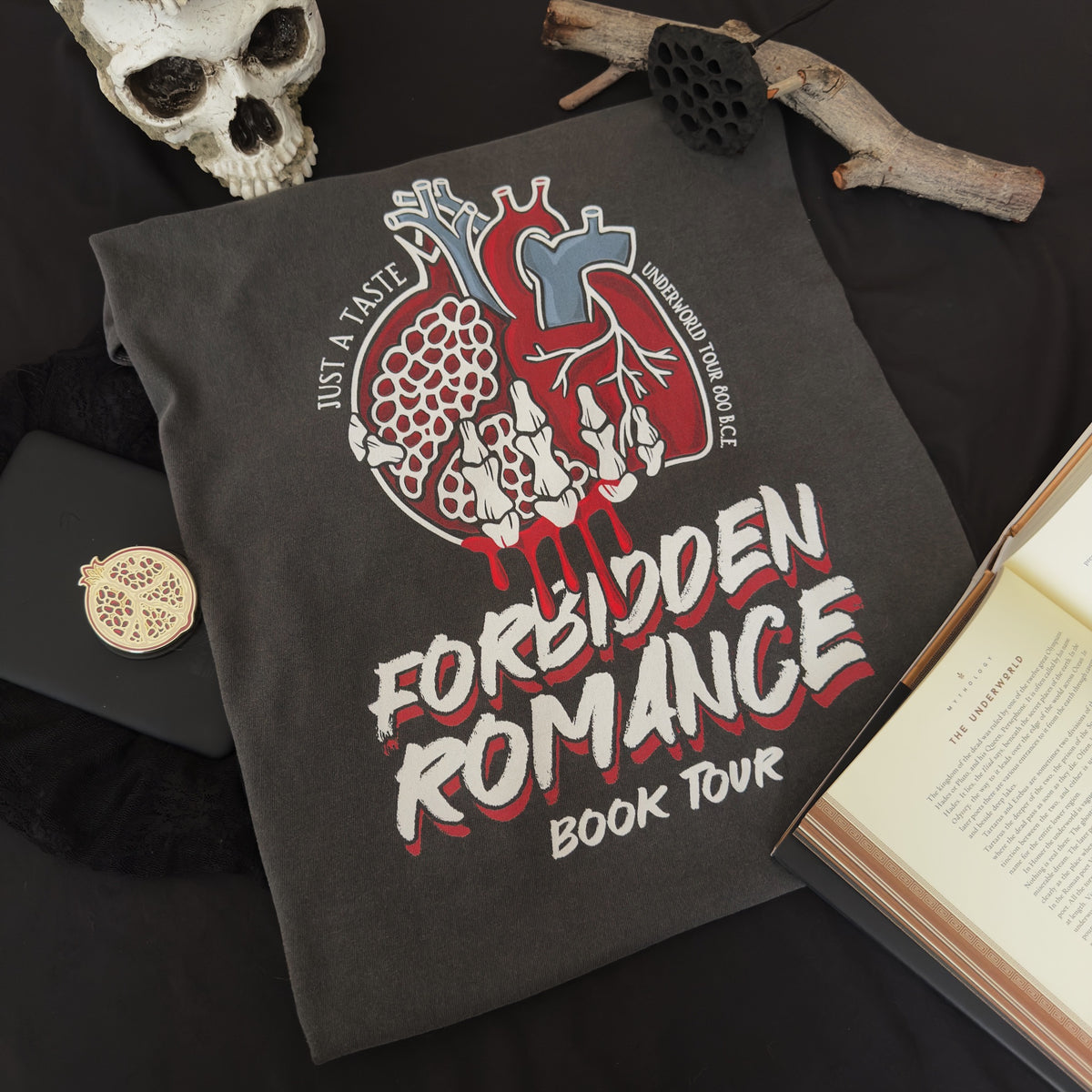 A dark charcoal gray graphic t-shirt laid flat on a black fabric background, surrounded by gothic-themed props including a skull, dried lotus pod, branch, and an open book. The t-shirt features a graphic of anatomical heart and pomegranate and a skeleton hand with the text 'JUST A TASTE,' 'UNDERWORLD TOUR 800 B.C.E.' and 'FORBIDDEN ROMANCE BOOK TOUR.'