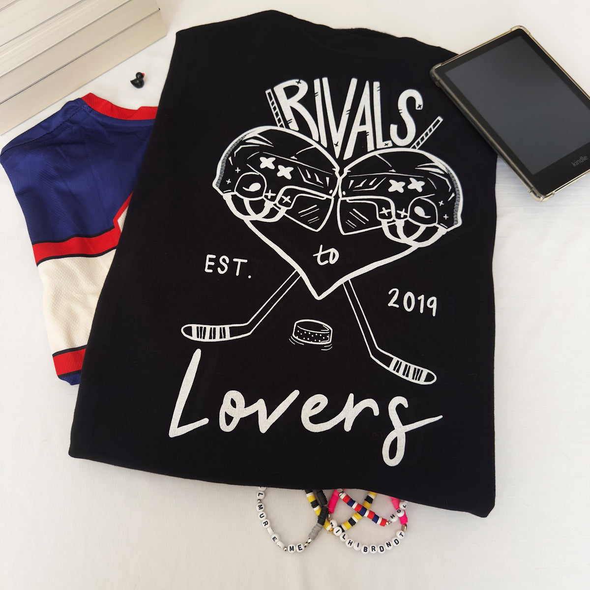 A black short-sleeve t-shirt laid flat on a white surface, featuring a white graphic print of two hockey helmets forming a heart, crossed hockey sticks, a hockey puck, and the text 'RIVALS to Lovers EST. 2019.'