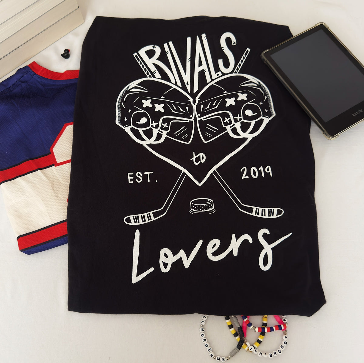 A black crewneck sweatshirt laid flat on a white surface, featuring a white graphic print of two hockey helmets forming a heart, crossed hockey sticks, a hockey puck, and the text 'RIVALS to Lovers EST. 2019.'