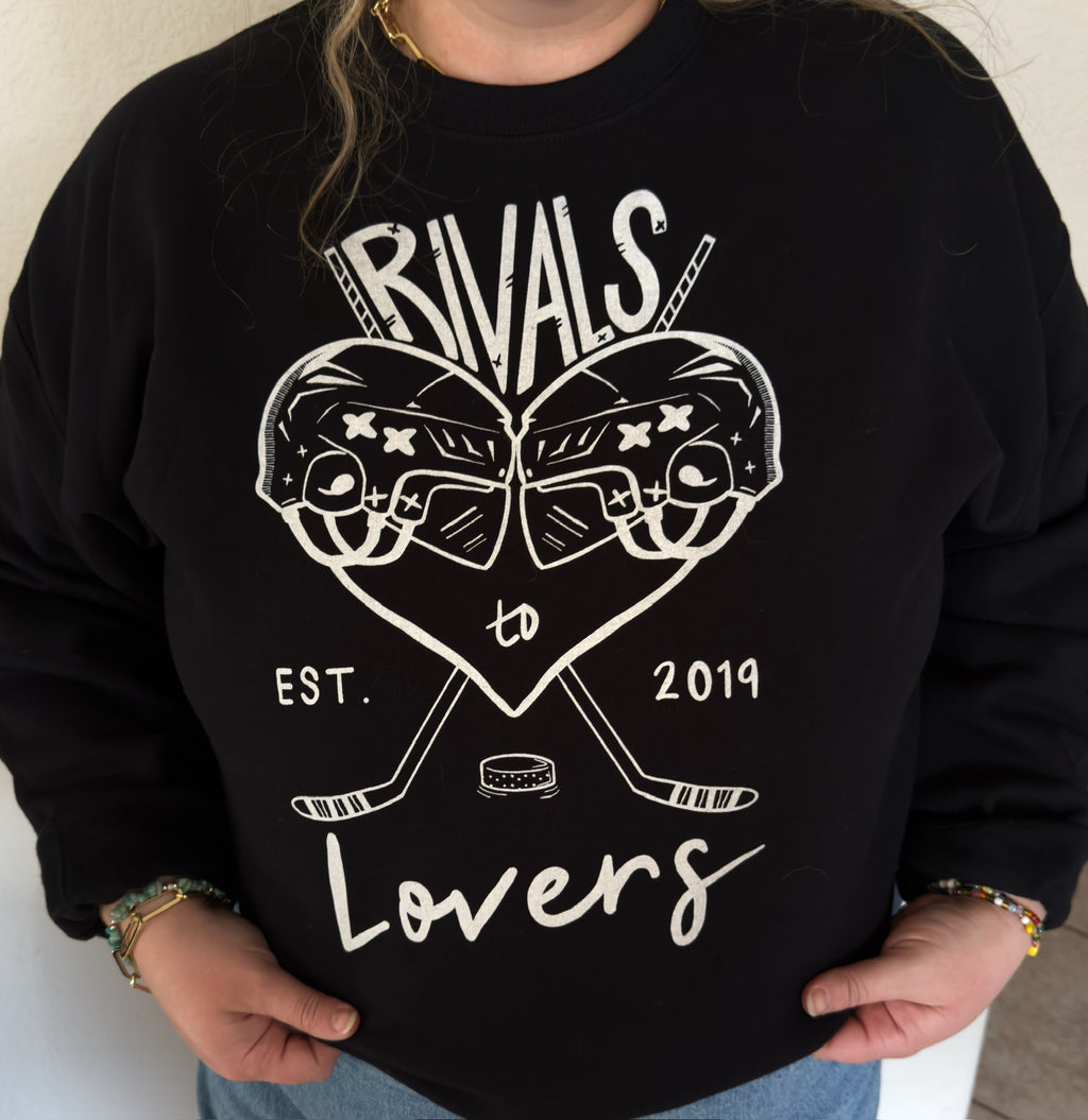 A person wearing a black crewneck sweatshirt, featuring a white graphic print of two hockey helmets forming a heart, crossed hockey sticks, a hockey puck, and the text 'RIVALS to Lovers EST. 2019.'