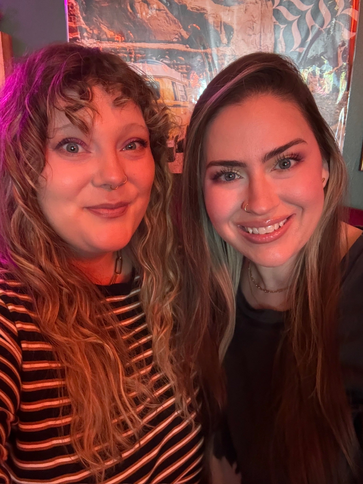 Two women smiling for a selfie in a dimly lit indoor setting with colorful ambient lighting. The woman on the left has wavy light brown blonde hair and wears a black and white striped shirt, while the woman on the right has long brown hair and is wearing a black top.